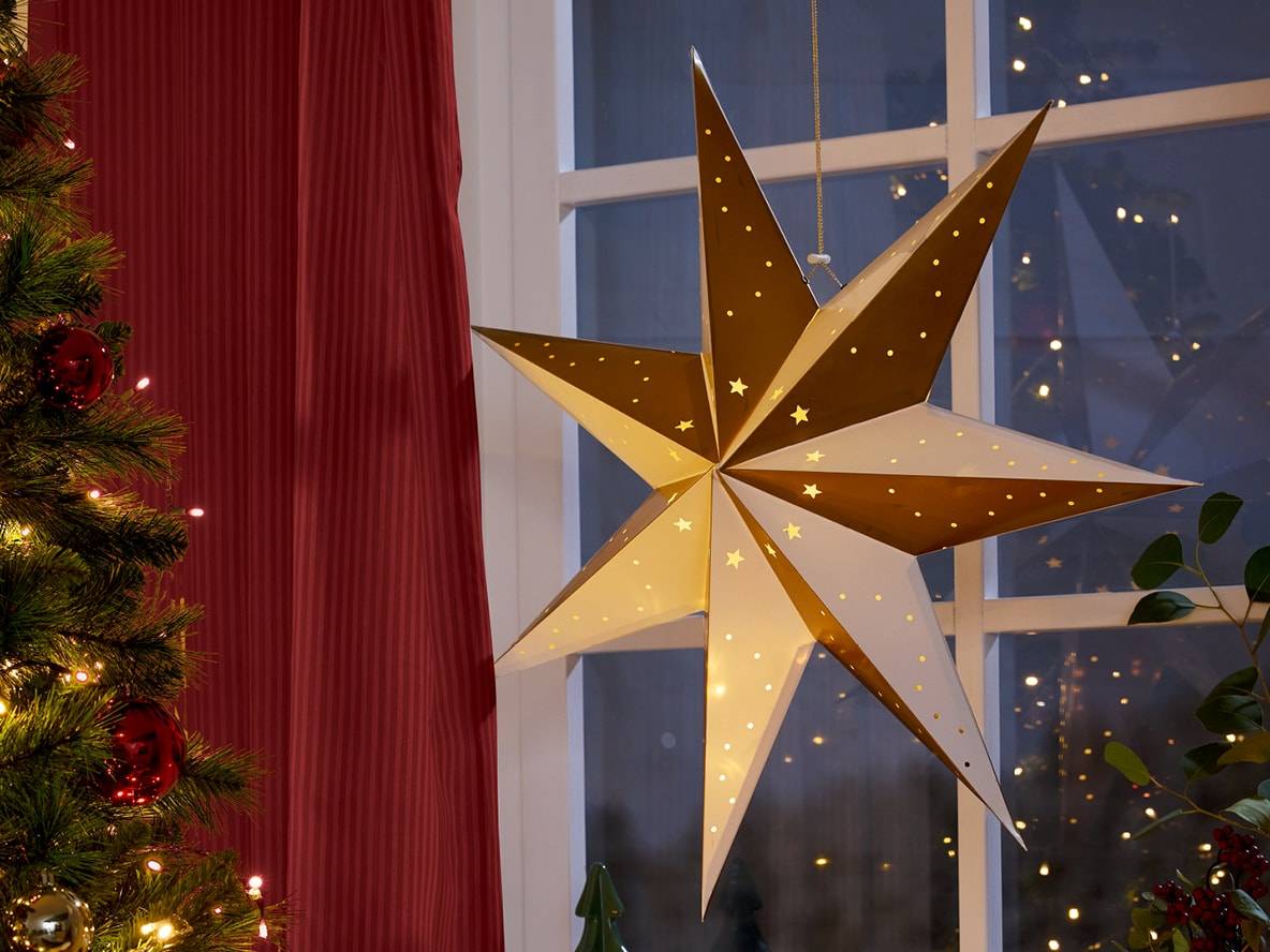 Leuchtender Weihnachtsstern hängt am Fenster, daneben ein Weihnachtsbaum und ein roter Vorhang.