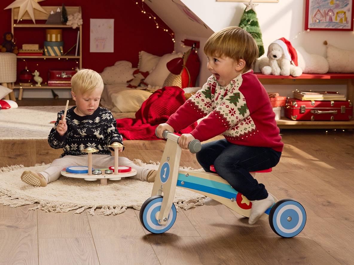 Zwei Kinder spielen mit einem Laufrad und einem Spielzeugschlagzeug in einem weihnachtlichen Zimmer.