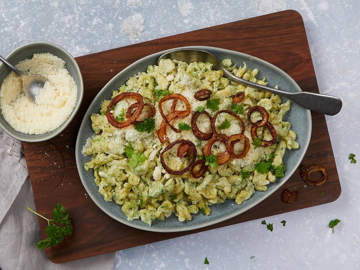 Grüne Spätzle mit Röstzwiebeln und geriebenem Parmesan auf einem Holzbrett.