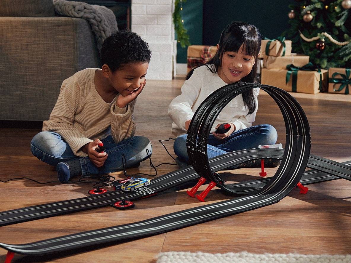 Deux enfants jouent avec un circuit de voitures Carrera sur un sol en bois.