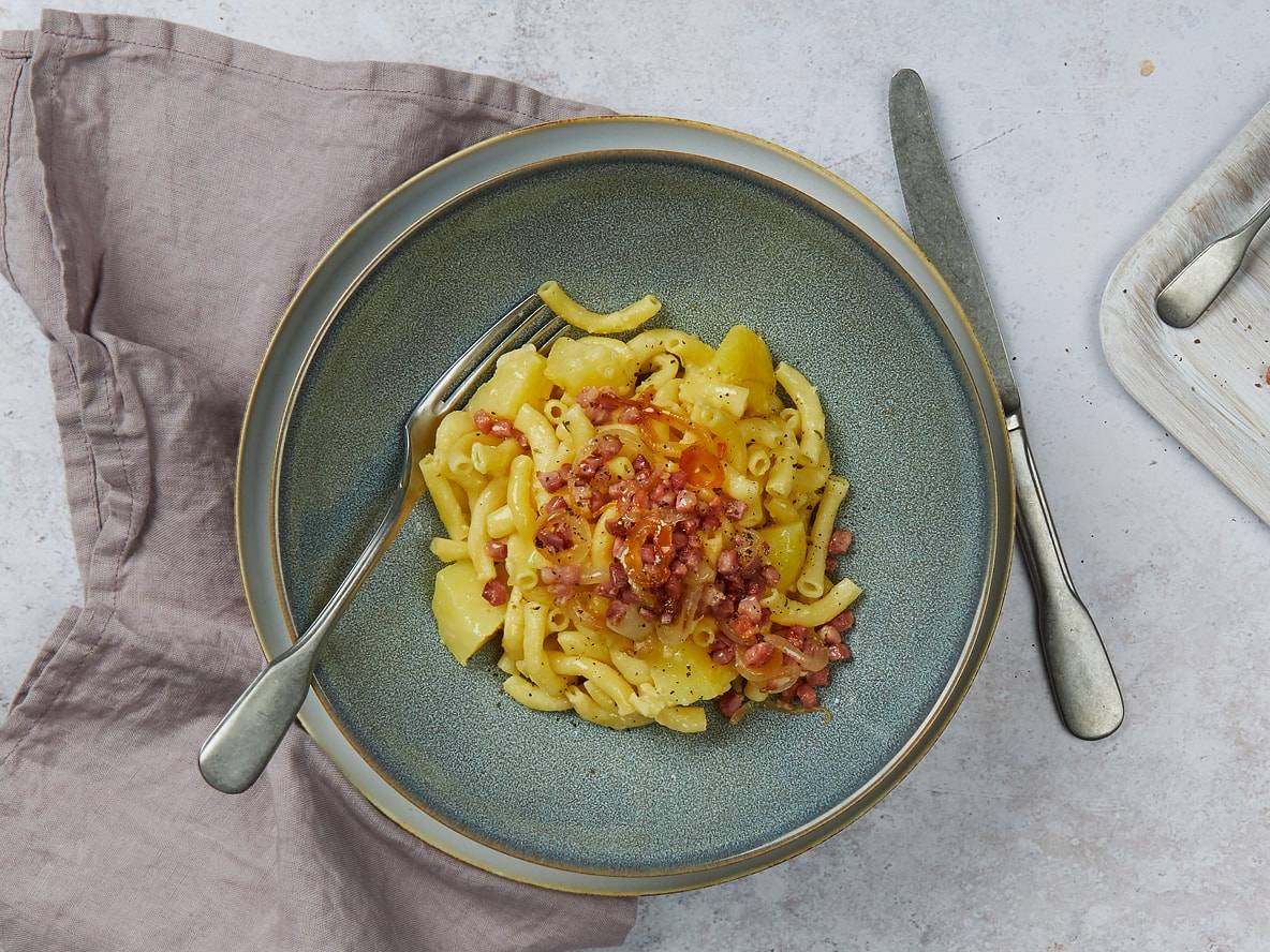 Makkaroni mit Kartoffeln und knusprigen Speckwürfeln in einer blauen Schale, mit Gabel und Messer.