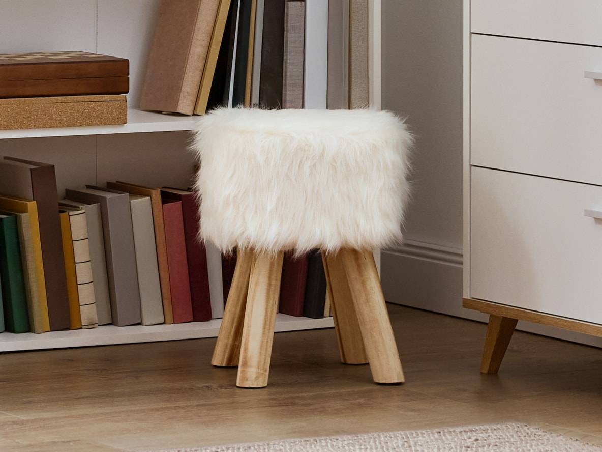Weißer Kunstfellhocker mit Holzbeinen, neben einem Bücherregal.