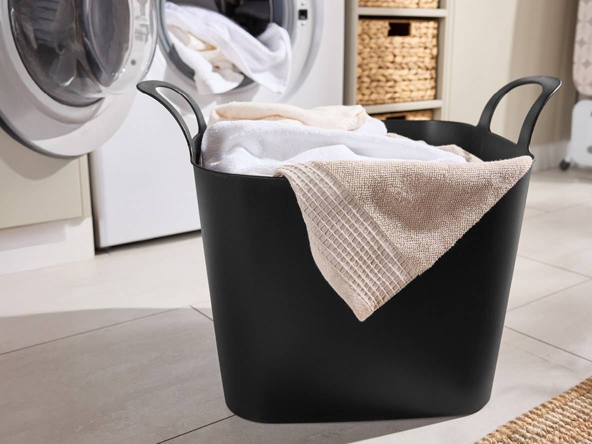 Schwarzer Wäschekorb gefüllt mit sauberen Handtüchern neben einer Waschmaschine.