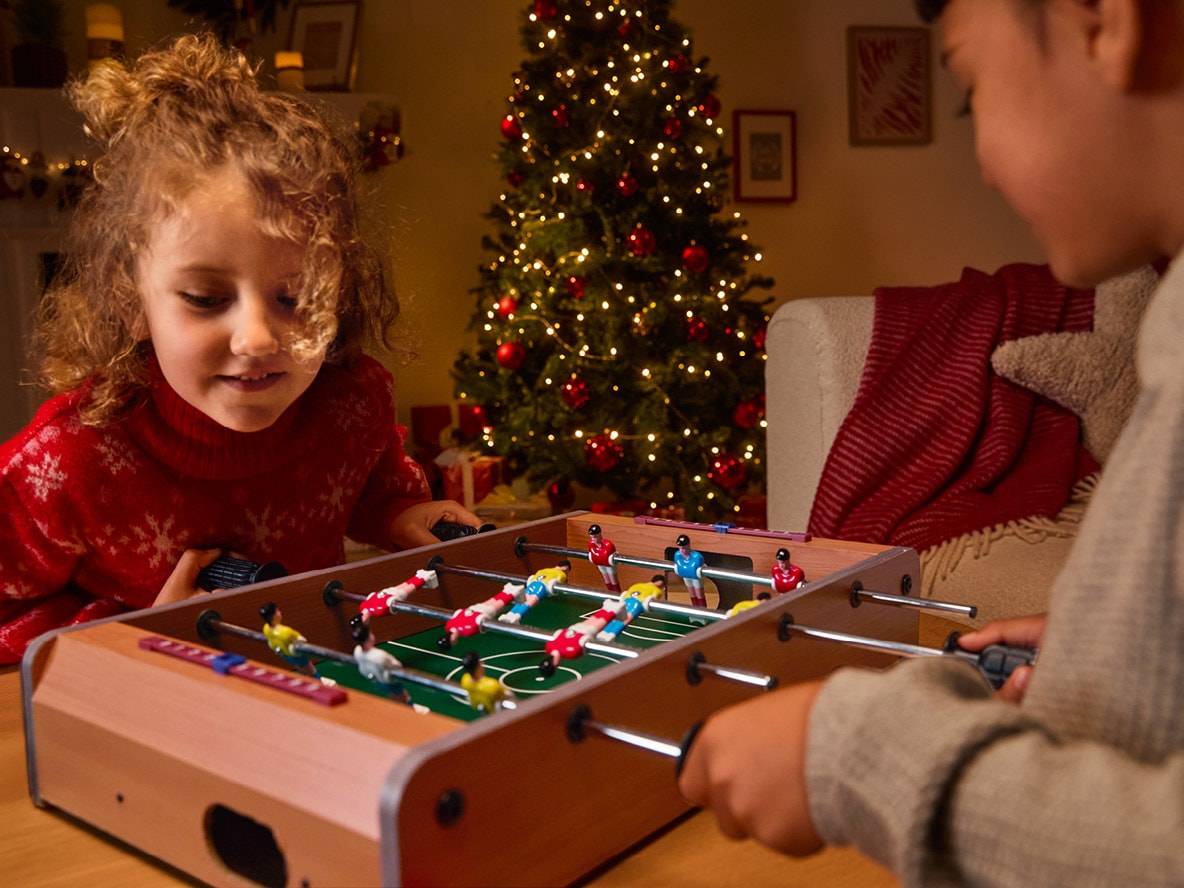 Zwei Kinder spielen Tischfußball vor einem beleuchteten Weihnachtsbaum.