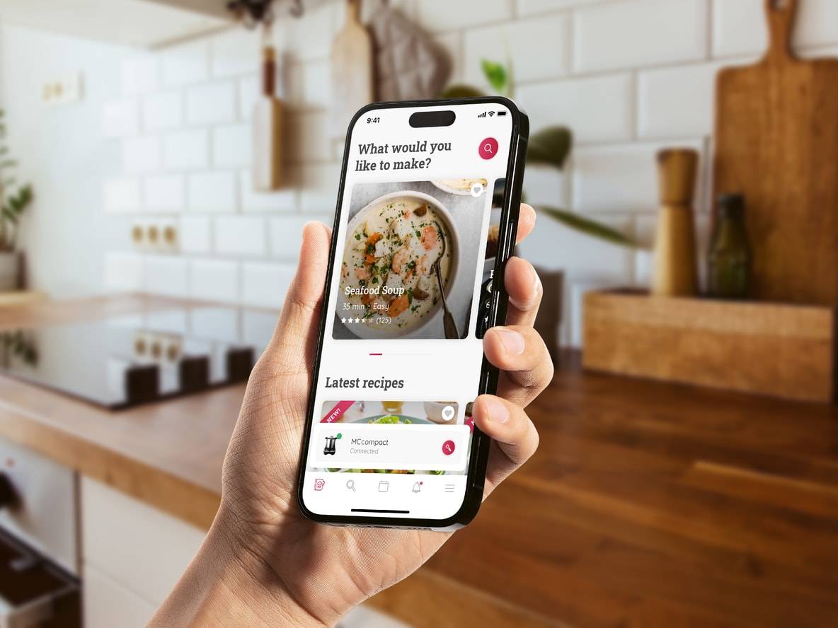 Eine Hand hält ein Smartphone mit einer Kochrezept-App in einer modernen Küche.
