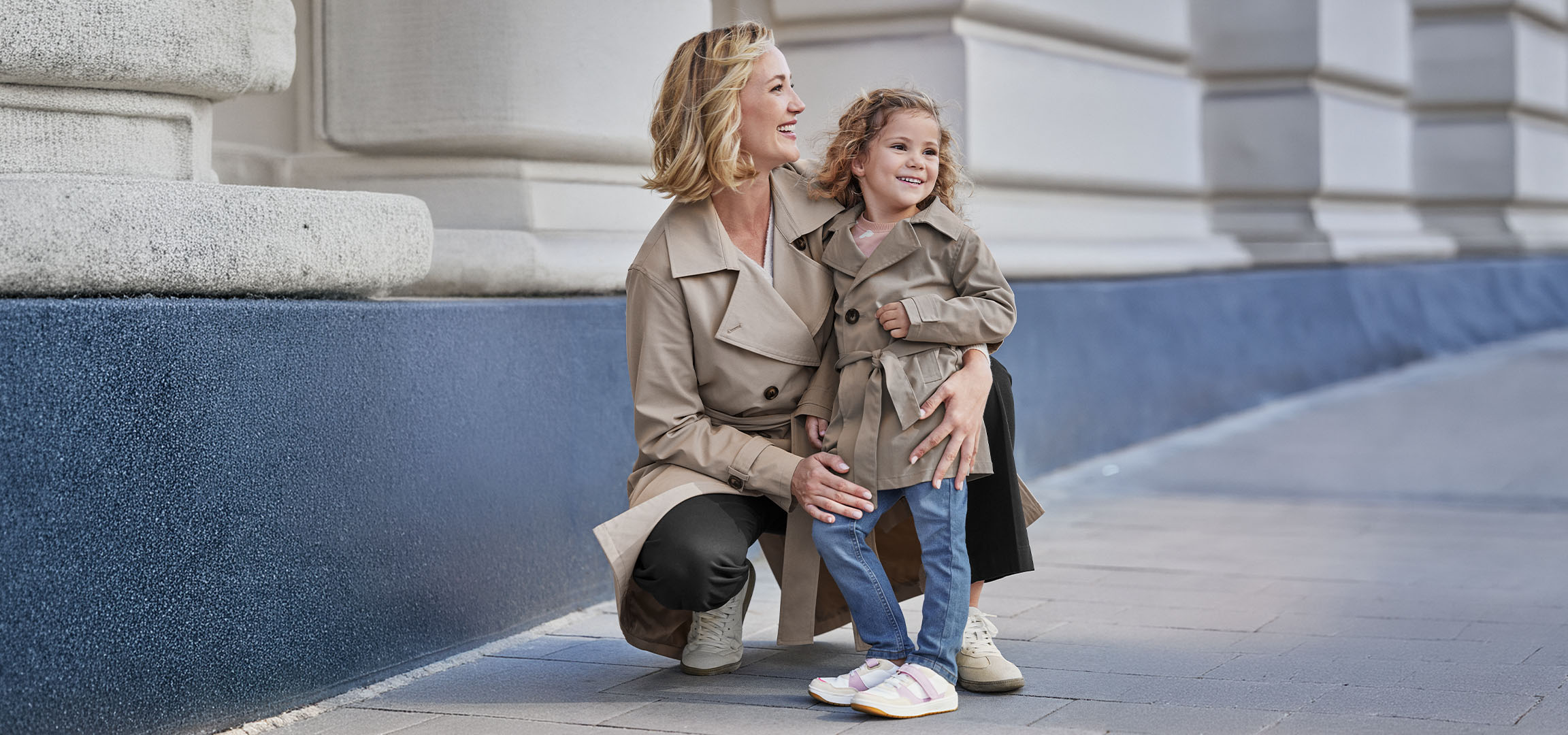 Deux femmes en trench-coats beiges, une adulte et une enfant, souriant à l'extérieur.