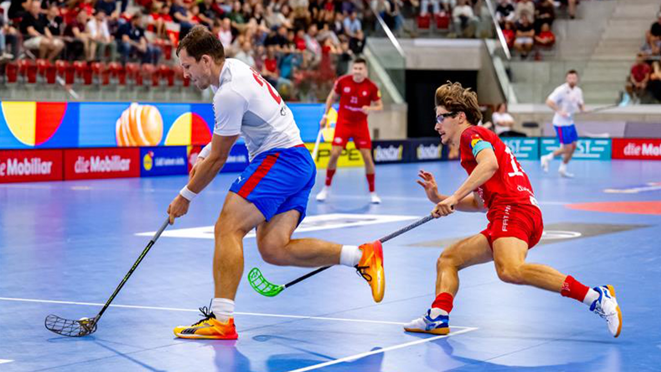 Due giocatori di floorball in azione su un campo blu, con uno che dribbla e l'altro che difende.