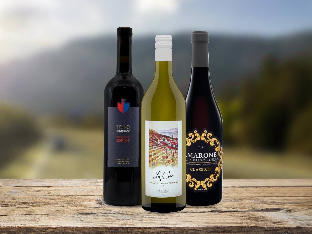 Trois bouteilles de vin : un rouge Ticino Merlot, un blanc La Côte et un rouge Marone Valpolicella Classico sur une table en bois.