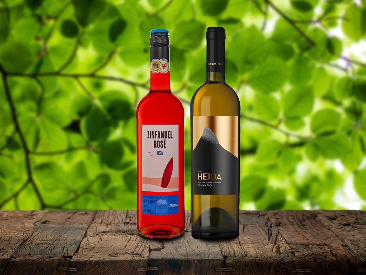 Zwei Weinflaschen, eine Rosé und eine Weißwein, auf einem Holztisch vor grünem Hintergrund.